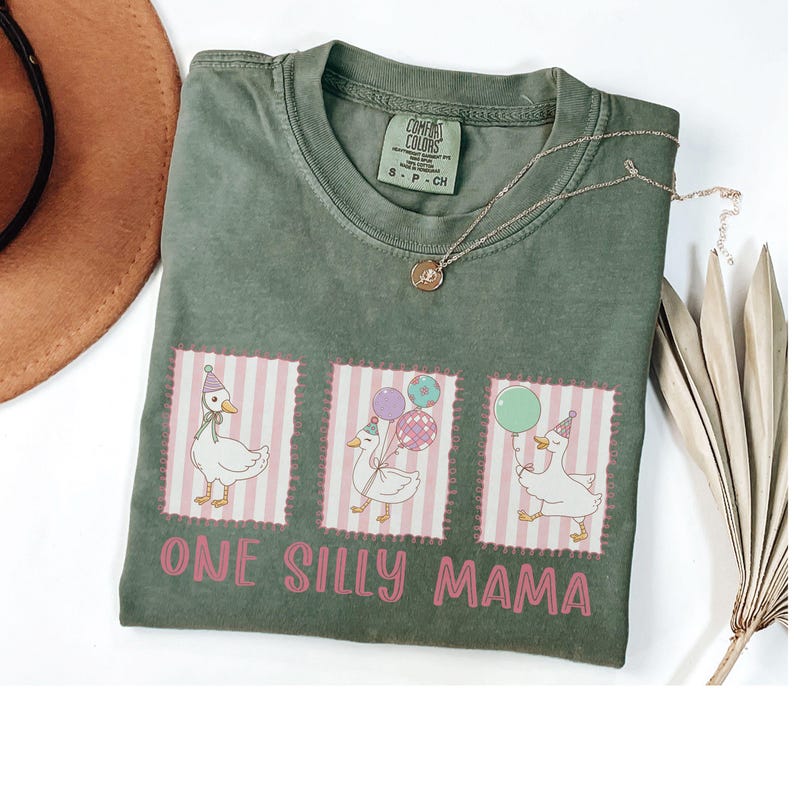 May include: Sage green t-shirt featuring the text "ONE SILLY MAMA" and three cartoon geese wearing party hats and holding balloons. The geese are in pink and white striped frames. A gold necklace is draped across the shirt.