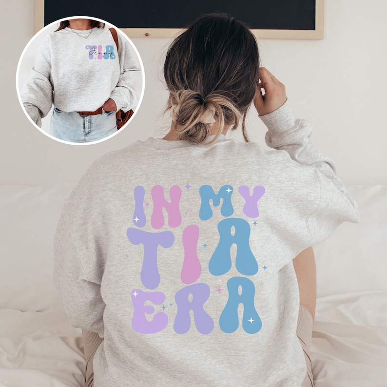 May include: Light gray sweatshirt with the text "In My Tia Era" in blue, purple, and pink bubble letters on the back. The front of the sweatshirt has the text "Tia" in a similar style. The sweatshirt is a casual, comfortable style.