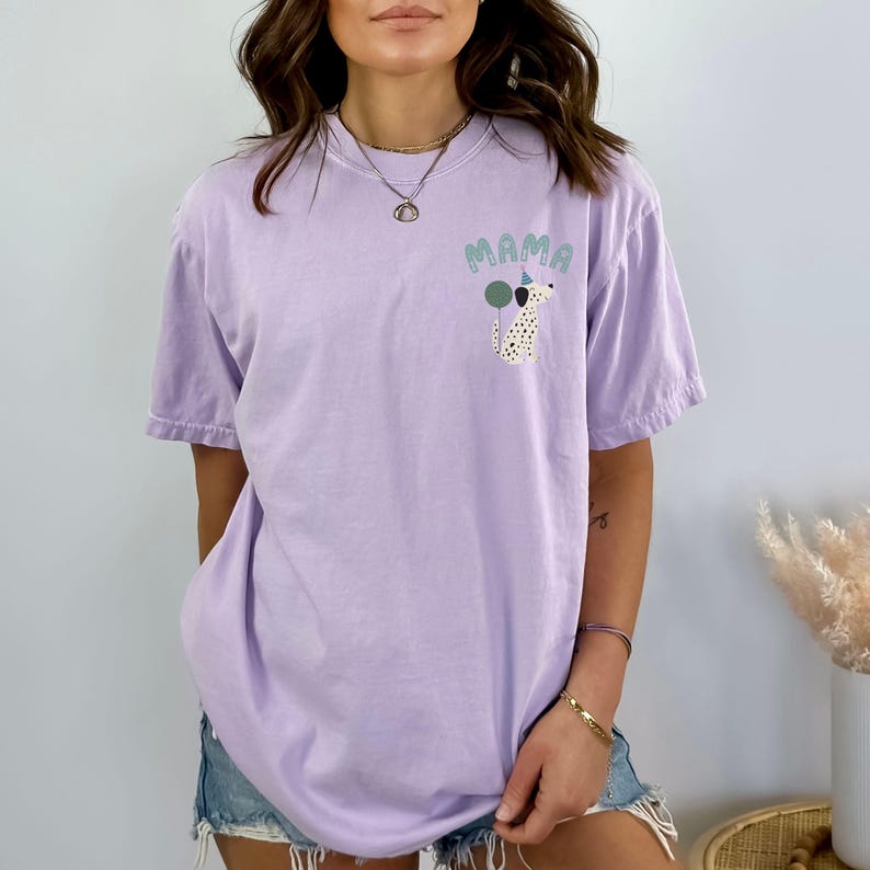 May include: A light purple t-shirt with a graphic of a Dalmatian dog wearing a party hat and holding a balloon. The word "MAMA" is above the dog in green letters. The shirt is a relaxed fit.