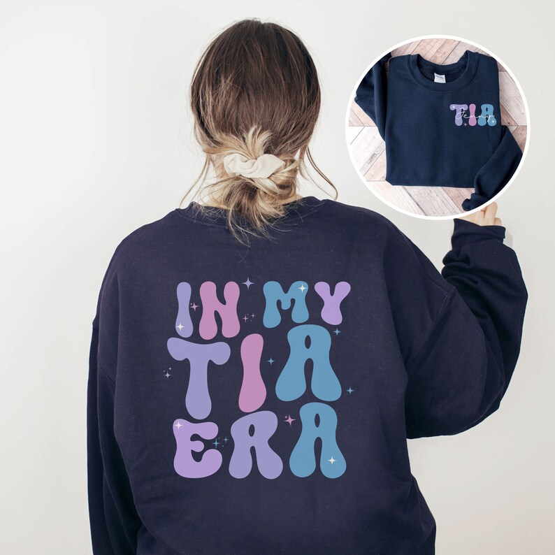 May include: Navy blue sweatshirt with the text "IN MY TIA ERA" in a retro font on the back. The front of the sweatshirt has the letters "TIA" in a similar style. The design features pastel colors.
