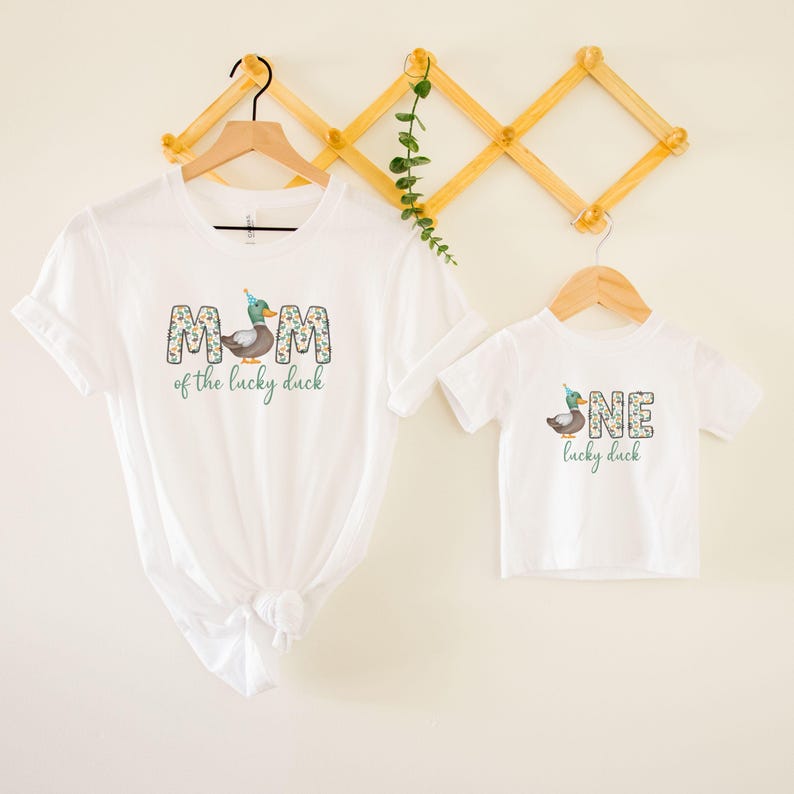Pode incluir: Duas camisetas brancas com um gr&aacute;fico de pato e texto. A camiseta maior diz "M&atilde;e do pato da sorte" e a camiseta menor diz "Um pato da sorte".