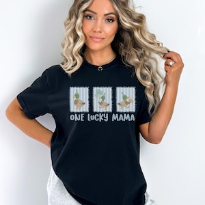 Puede incluir: Camiseta negra con un gr&aacute;fico de tres patos con sombreros de fiesta, sobre un fondo a rayas azules y blancas. El texto "ONE LUCKY MAMA" est&aacute; impreso debajo del gr&aacute;fico. La camiseta es de estilo casual.