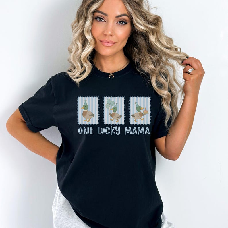 Puede incluir: Camiseta negra con un gr&aacute;fico de tres patos con sombreros de fiesta, sobre un fondo a rayas azules y blancas. El texto "ONE LUCKY MAMA" est&aacute; impreso debajo del gr&aacute;fico. La camiseta es de estilo casual.