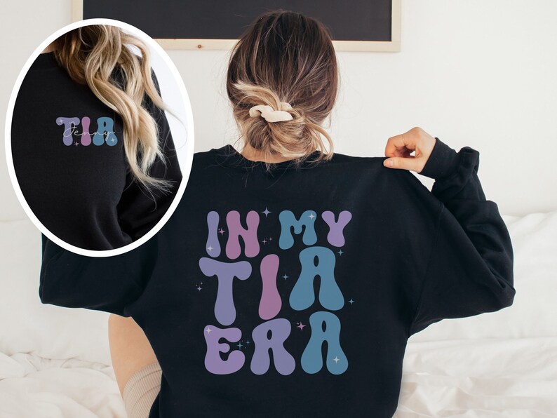 May include: Black sweatshirt with the text "IN MY TIA ERA" in a retro font on the back. The front left shoulder has the word "TIA" in a similar font. The letters are in shades of purple and blue with star accents.