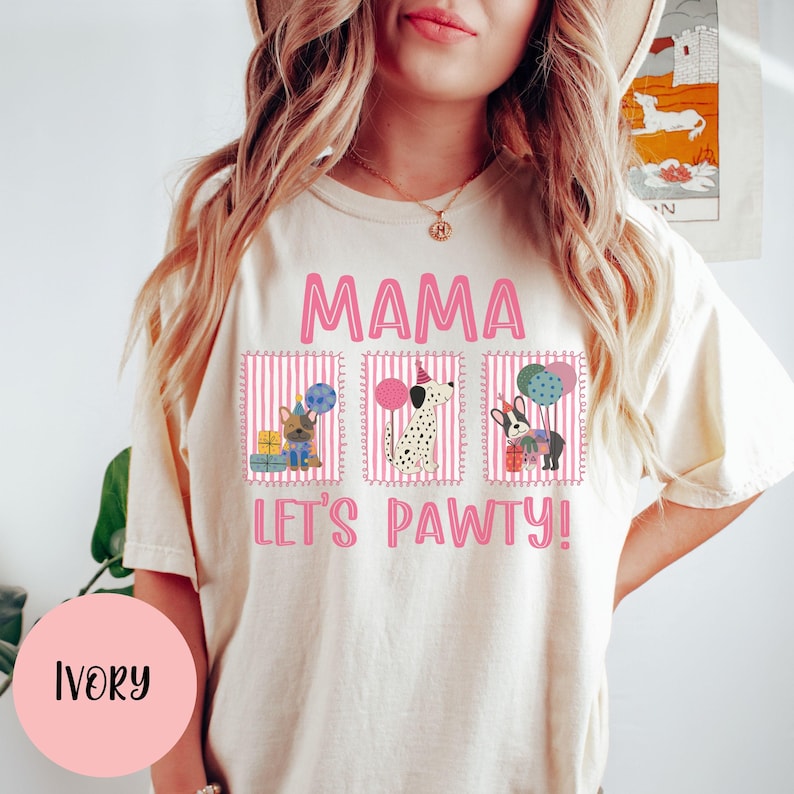 May include: Ivory-colored t-shirt with the text "MAMA LET'S PAWTY!" in pink. The design features three dog illustrations with party hats and balloons. The shirt is a casual, comfortable style.