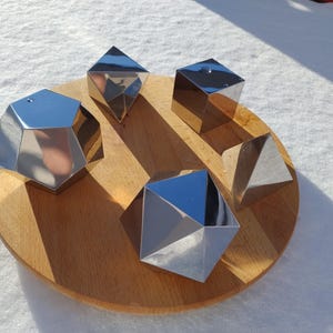 May include: Five shiny, silver geometric shapes, including a cube, a pyramid, and three irregular polyhedrons, are arranged on a circular wooden tray.