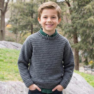 May include: A grey, hand-knitted jumper with a crew neck. The jumper is worn over a green collared shirt and paired with blue jeans. The person is smiling with their hands in their pockets. The background features trees and a large rock.