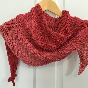 May include: A coral-coloured, hand-knitted shawl with a triangular shape. The shawl features a textured stitch pattern and is displayed on a wooden hanger. The knitted design includes openwork details and a fringed edge.