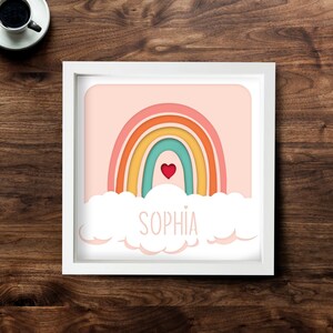 May include: A framed art print featuring a colorful rainbow design with a heart and the name "Sophia" in a playful font. The rainbow has layers of orange, yellow, teal, and pink. The print is set against a light pink background.