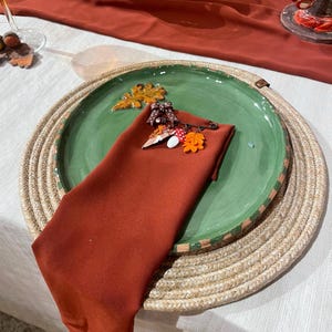 Puede incluir: Un plato redondo de cerámica verde con relieve de hoja de roble, cubierto con una servilleta de tela color óxido doblada. Un broche decorativo de metal con un lazo marrón de lunares, un pequeño hongo blanco y una hoja naranja sujeta la servilleta.