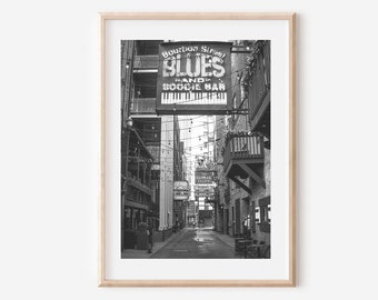 Puede incluir: Fotografía en blanco y negro de un callejón estrecho en Nueva Orleans, Luisiana. Un letrero sobre la entrada dice "Bourbon Street Blues Band and Boogie Bar" con un gráfico de teclado de piano. Luces de cadena cuelgan arriba.