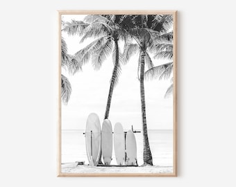 May include: Black and white photograph of four surfboards leaning against a palm tree on a beach. The surfboards are white and the palm tree has lush green leaves.