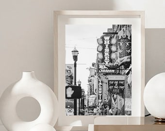 Puede incluir: Impresión enmarcada en blanco y negro de una escena callejera con letreros de neón, incluyendo "Whiskey Row Saloon". Un jarrón blanco con una abertura circular y una pequeña esfera blanca están en primer plano.