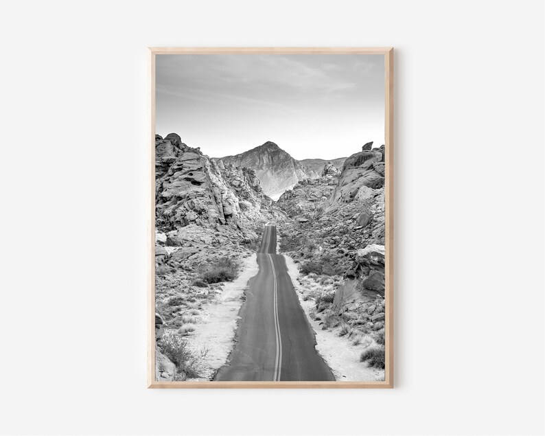 May include: A black and white photograph of a winding road through a desert landscape. The road is lined with rocky hills and mountains in the distance.