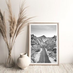 May include: Black and white photograph of a winding road through a desert landscape with mountains in the background. The photo is framed in a light wood frame.