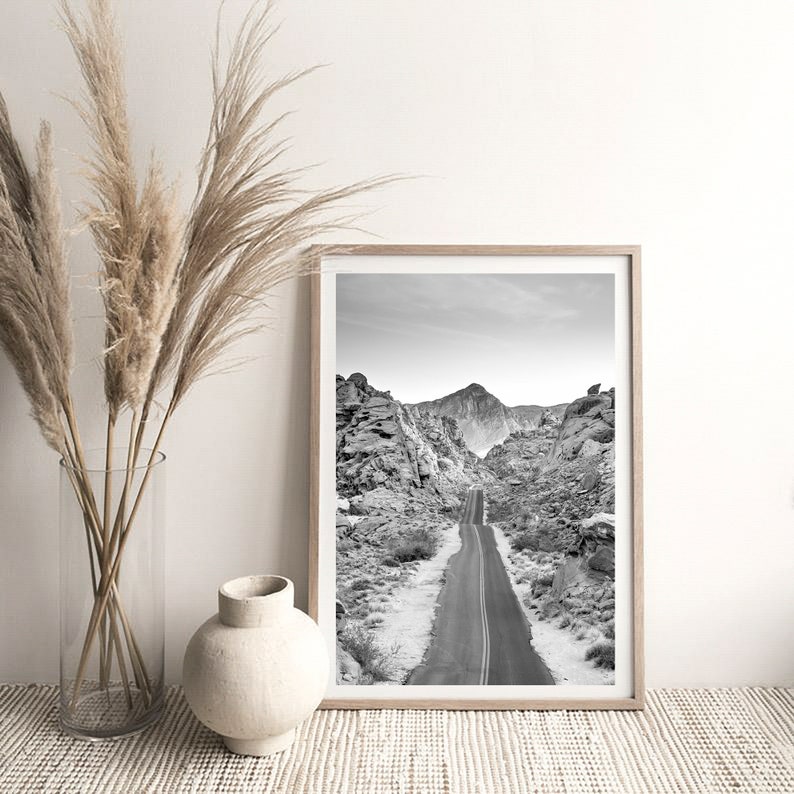 May include: Black and white photograph of a winding road through a desert landscape with mountains in the background. The photo is framed in a light wood frame.