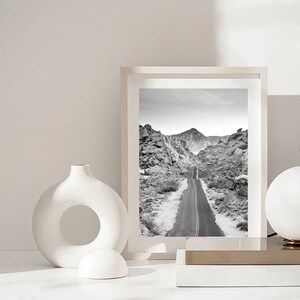 May include: A black and white photograph of a road winding through a desert landscape. The photo is framed in a white frame and is displayed on a white surface with a white ceramic vase and a white ceramic sphere.