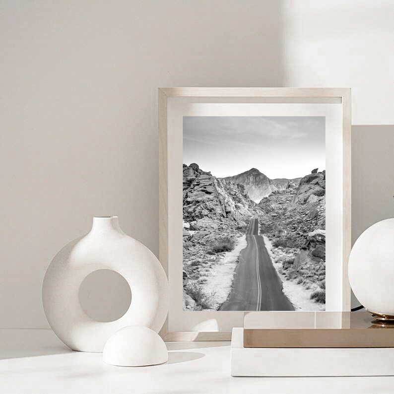 May include: A black and white photograph of a road winding through a desert landscape. The photo is framed in a white frame and is displayed on a white surface with a white ceramic vase and a white ceramic sphere.