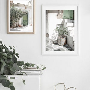 May include: Two framed photographs of whitewashed buildings with green doors and potted plants in a white room with a white cabinet and a woven basket.