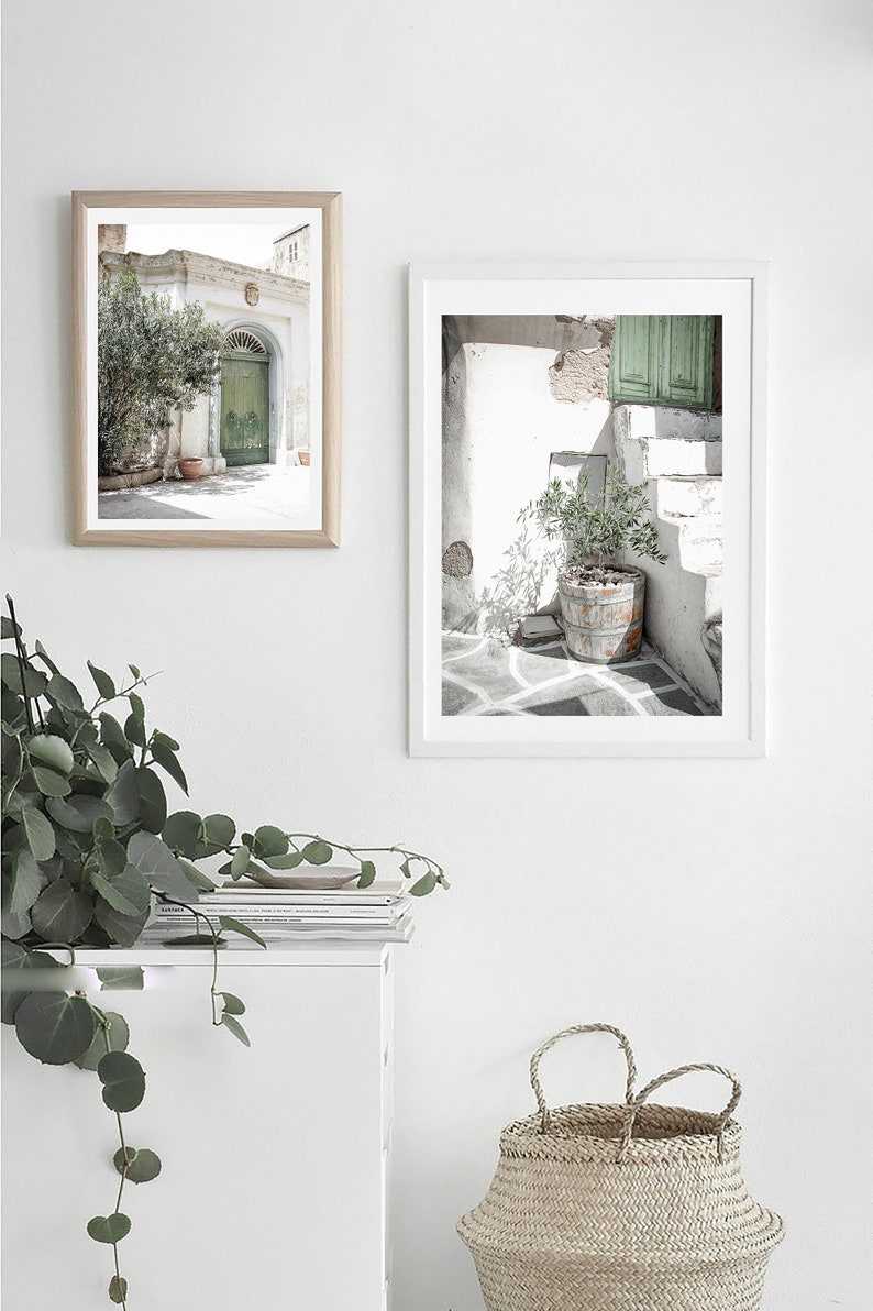 May include: Two framed photographs of whitewashed buildings with green doors and potted plants in a white room with a white cabinet and a woven basket.