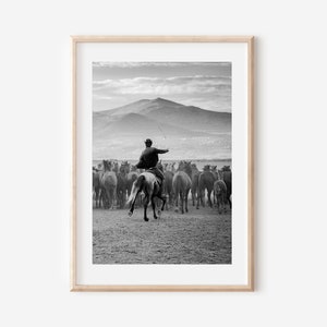 May include: Black and white framed print of a cowboy on horseback herding a large group of horses. The cowboy is wearing a hat and jacket, and is holding a whip. Mountains are visible in the background.