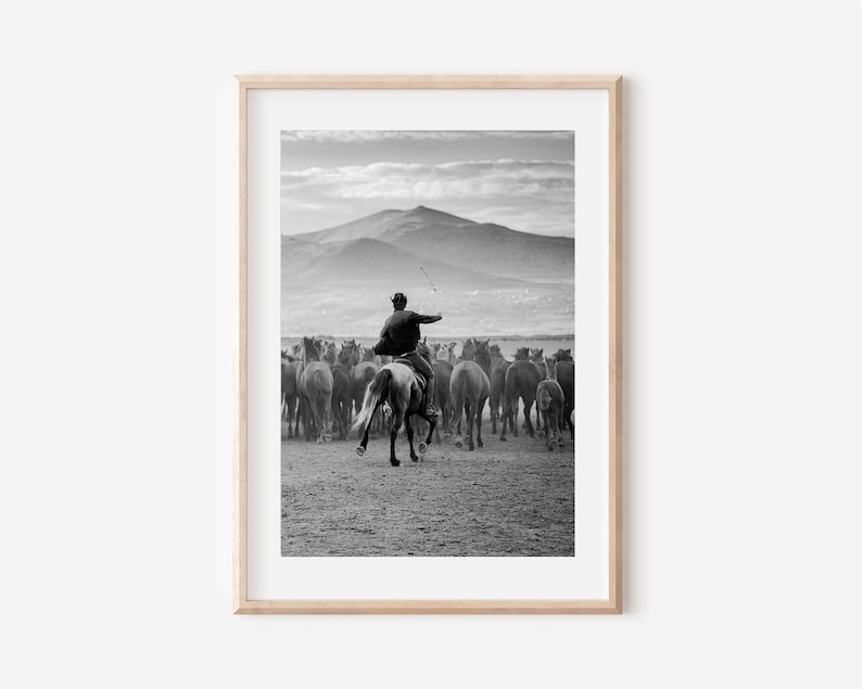 May include: Black and white framed print of a cowboy on horseback herding a large group of horses. The cowboy is wearing a hat and jacket, and is holding a whip. Mountains are visible in the background.