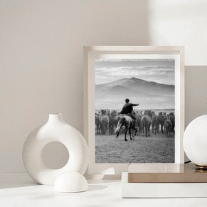 May include: Black and white framed print of a cowboy on horseback herding a group of horses across a field. The background features a mountain range and a cloudy sky. The print is displayed on a white surface.