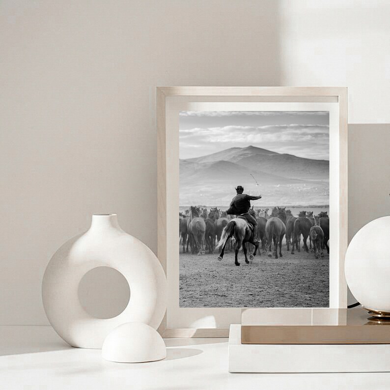 May include: Black and white framed print of a cowboy on horseback herding a group of horses across a field. The background features a mountain range and a cloudy sky. The print is displayed on a white surface.