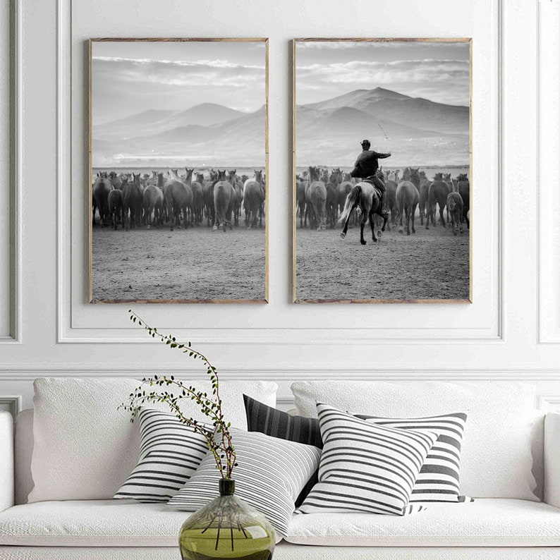 May include: A set of two framed black and white prints. One shows a herd of horses, the other a rider on horseback herding the horses. The background features mountains and a cloudy sky. The prints are displayed above a white sofa with decorative pillows.