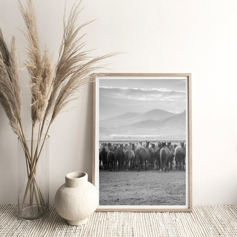 May include: Black and white framed print featuring a herd of horses in a field, with mountains in the background. The print is displayed on a white surface, next to a vase with tall, dried grasses and a white ceramic vase.