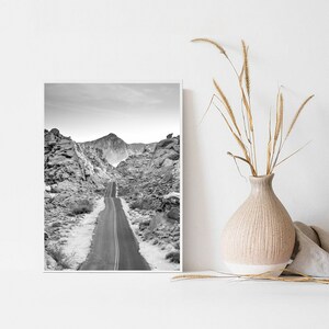 May include: Black and white photograph of a winding road through a desert landscape with mountains in the background.