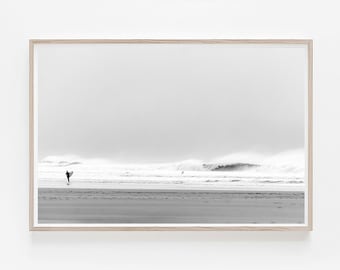 Peut inclure: Une photographie en noir et blanc d'un surfeur marchant sur une plage avec une planche de surf sous le bras. L'océan est en arrière-plan avec des vagues qui se brisent.