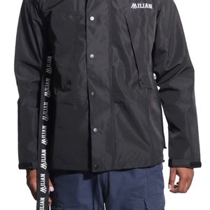 May include: A black hooded jacket with a zip and button fastening. The jacket has a white logo that says "MILLIAN" on the right side of the chest. The jacket has a white "MILLIAN" logo repeated on the left side of the jacket.