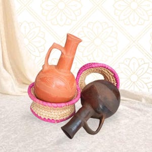 Peut inclure: Deux cruches en argile faites à la main, une orange et une marron foncé, sont exposées. La cruche orange est posée sur un panier tressé avec une bordure rose. Un deuxième panier tressé et une cruche marron foncé sont également visibles. Le fond présente un motif floral.