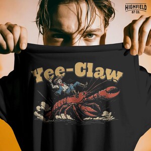 Crawfish rodeo humor design featuring a cowboy riding a large crawdad while dust flies behind the claws. The Yee Claw phrase appears above the scene making this a playful Cajun themed artwork.