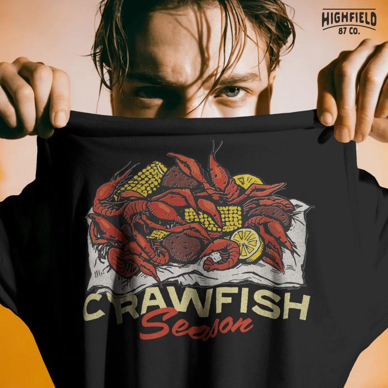 Seafood boil themed shirt design featuring a pile of crawfish with lemon slices and corn on paper. The bold Crawfish Season text emphasizes summer gatherings and southern food culture.