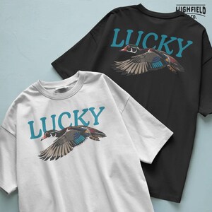 Waterfowl PNG illustration featuring a wood duck flying across the design with teal Lucky text behind it. The artwork uses bold outlines, flat colors, and detailed feather shapes for bird lover apparel, outdoor graphics, nature shirts, and digital download projects.