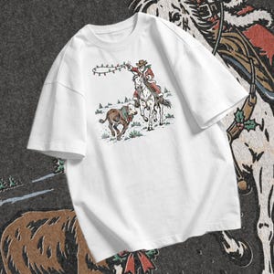 Cowboy Christmas design featuring Santa riding a white horse beside a festive calf with evergreen greenery, red berries, and holiday ribbon.