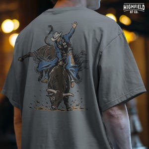 Rodeo shirt PNG showing a bull rider gripping the rope while the bull bucks forward, ideal for western clothing and cowboy lifestyle designs.