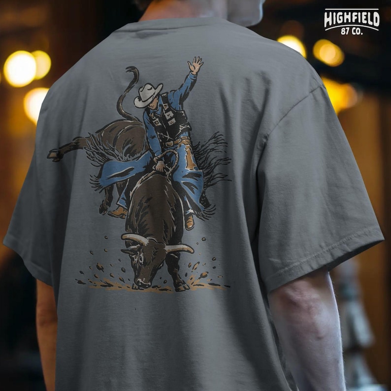 Rodeo shirt PNG showing a bull rider gripping the rope while the bull bucks forward, ideal for western clothing and cowboy lifestyle designs.