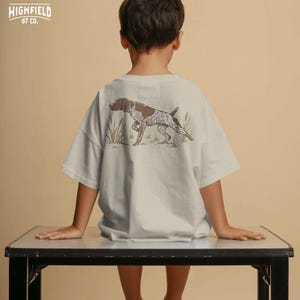 Hunting shirt PNG showing a German Shorthaired Pointer bird dog pointing in grass, designed for upland hunters and outdoorsmen.
