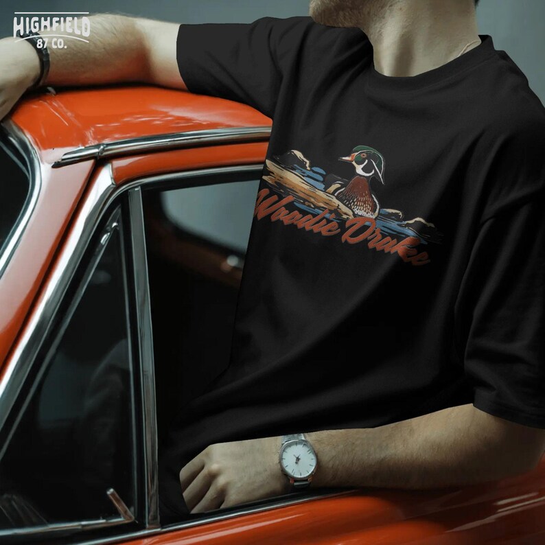 Vintage waterfowl PNG featuring a wood duck drake in a pond scene with Woodie Drake text, classic hunting illustration