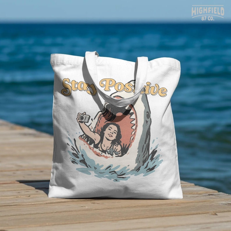 humorous beach scene with shark and woman taking selfie inside mouth, stay positive phrase, funny ocean png design for summer shirts and digital use