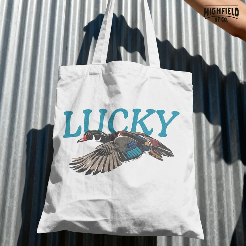 Lucky Duck digital download artwork featuring a colorful wood duck in flight with teal lettering, detailed wings, and a clean white background. This PNG is made for shirt makers, bird lovers, outdoor style apparel, and waterfowl themed products.