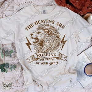 May include: A white t-shirt with a brown lion graphic and the text "The Heavens Are Roaring The Praise Of Your Glory".