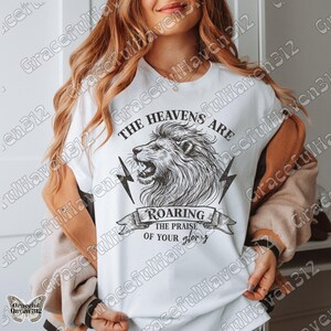 May include: White t-shirt with a black and white illustration of a lion's head roaring. The text "The Heavens Are Roaring The Praise Of Your Glory" is printed on the shirt.