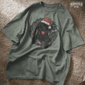 Happy black lab in Santa hat surrounded by pine leaves and holiday decor, including a bone and a pink whale toy. PNG format for seasonal crafting.