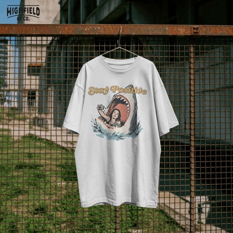 shark artwork with woman smiling inside shark mouth holding camera, stay positive text, funny beach png design for vacation shirts and ocean humor apparel