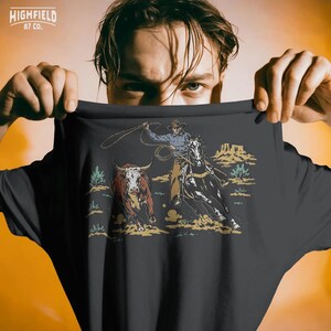 Team roping shirt design with cowboy, black horse, running steer, rope loop, sagebrush, and western ranch style artwork.