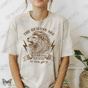 May include: A beige t-shirt with a brown graphic of a roaring lion with lightning bolts and the text "The Heavens Are Roaring The Praise Of Your Glory".